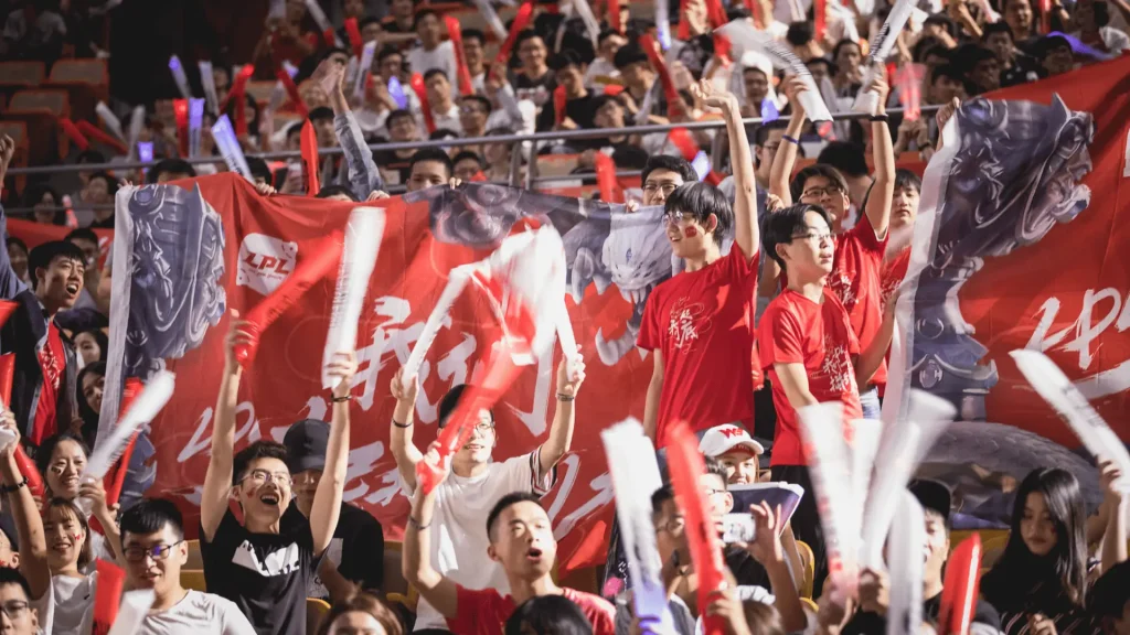 Crowd of LPL esports fans cheering with red banners, clappers, and team flags in packed arena stands